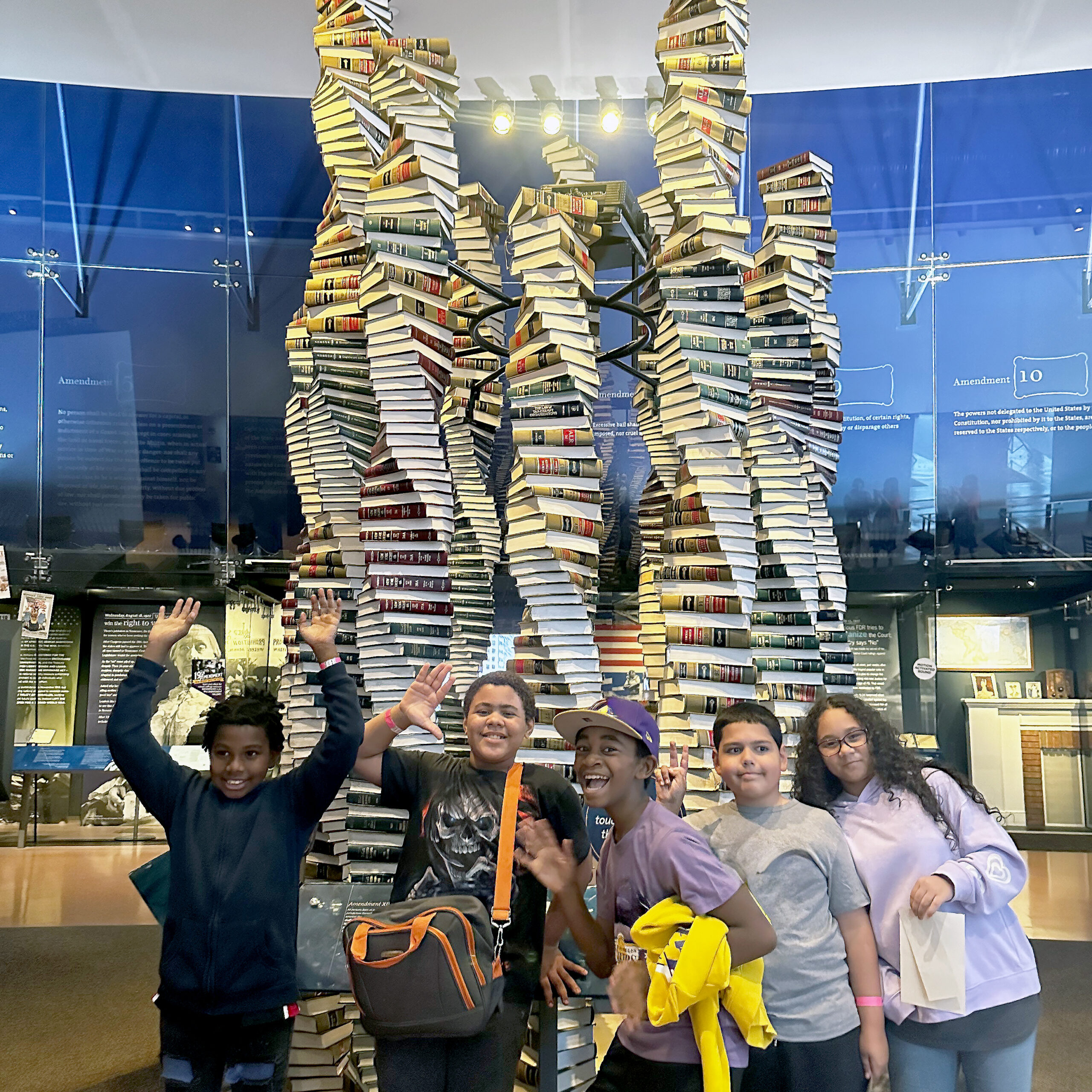 Students pose and wave in front of a large sculpture made of stacked books shaped like DNA strands.