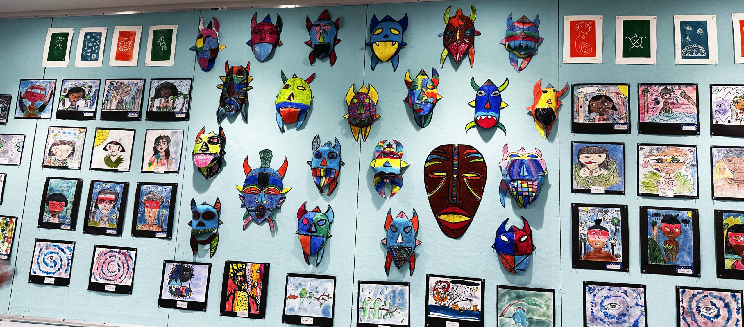 Colorful student artwork and 3D masks displayed on a blue wall in a school or gallery setting.